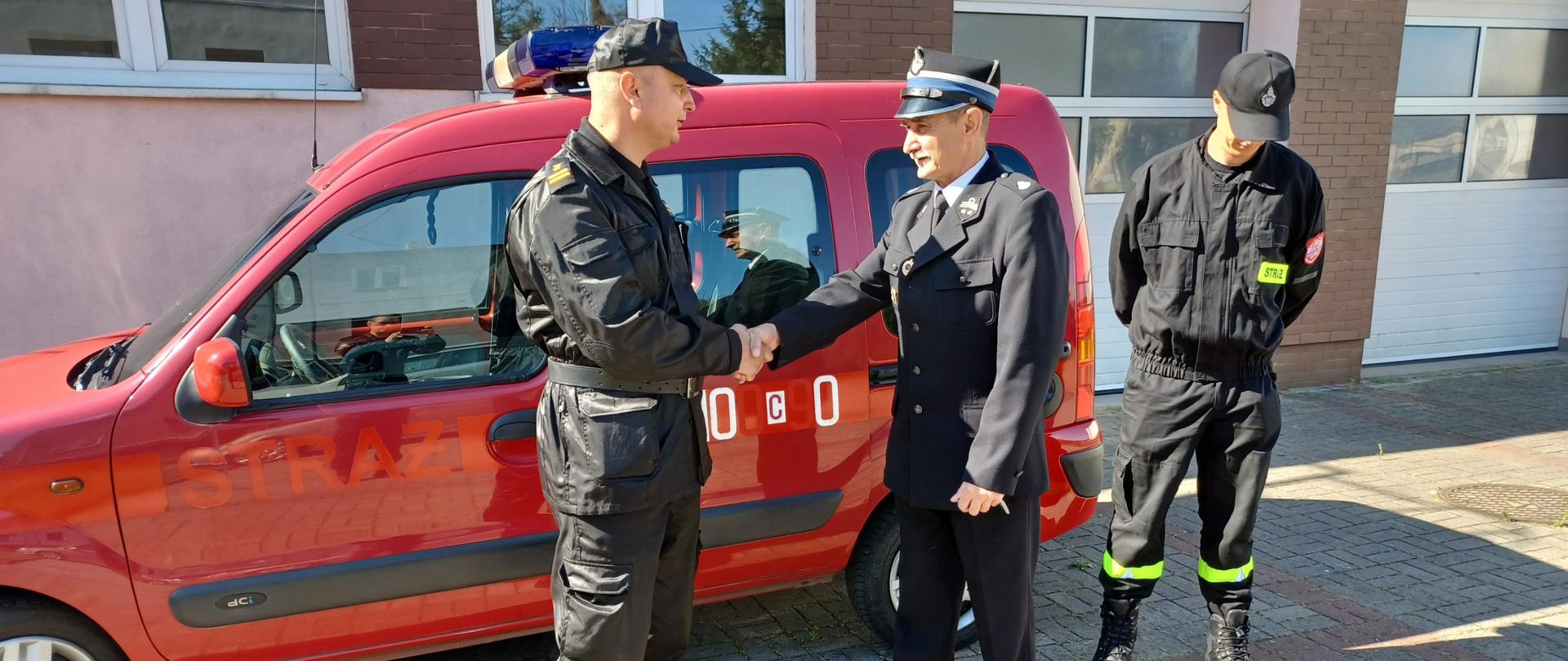 Zdjęcie obrazuje trzech mężczyzn stojących na tle samochodu osobowego w kolorze czerwonym marki Renault. Dwóch z nich uściska sobie dłoń. Jeden z nich jest ubrany w umundurowanie galowe koloru granatowego oraz ma założoną na głowie czapkę rogatywkę koloru granatowego z czarnym daszkiem i emblematami Ochotniczej Straży Pożarnej, natomiast drugi z nich ubrany jest w czarne umundurowanie dwu częściowe z zapiętym skórzanym pasem oraz założoną na głowie czarną czapką typu bejsbolową. Lekko po prawej stronie stoi jeszcze trzeci mężczyzna, funkcjonariusz ochotniczej straży pożarnej w umundurowaniu tak zwanym koszarowym złożonym z dwóch części w kolorze czarnym z żółtymi odblaskami na spodniach oraz na piersi. Za mężczyznami stoi czerwony samochód marki Renault z niebieskimi światłami sygnałowymi na dachu oraz srebrnymi kołpakami na kołach. W tle za samochodem widać budynek garażowy z zamkniętymi bramami w kolorze białym, budynek jest koloru wyblakniętego różu oraz na elewacji widać brązowe ozdobne cegły klinkierowe.