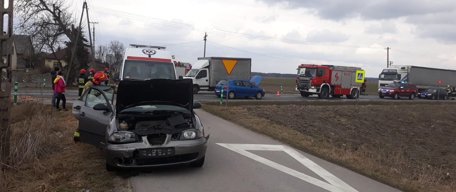Zdjęcie przedstawia na pierwszym planie przód uszkodzonego pojazdu osobowego w kolorze szarym. W samochodzie otwarta jest maska silnika oraz drzwi po prawej stronie. Na drugim planie znajduje się drugi uszkodzony pojazd, który brał udział w zdarzeniu oraz dwa samochody strażackie. Po drodze wahadłowo poruszają się inne pojazdy. Po lewej stronie widać kilku strażaków w ubraniu specjalnym oraz kilku świadków zdarzenia. 