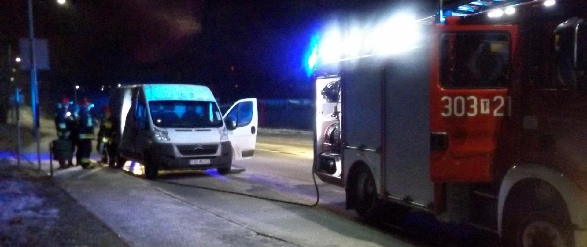 Na zdjęciu znajduje się samochód gaśniczy na sygnałach. Jest ciemno. Za samochodem gaśniczym stoi bus przewożący pieczywo. Przed nim strażacy sprawdzający miejsce pożaru.