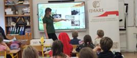 Inspektorka IJHARS prowadzi lekcję w Szkole Podstawowej nr 51 – stoi przy tablicy, a w tle widoczny jest roll-up z logo IJHARS; uczniowie siedzą w ławkach i uważnie słuchają.