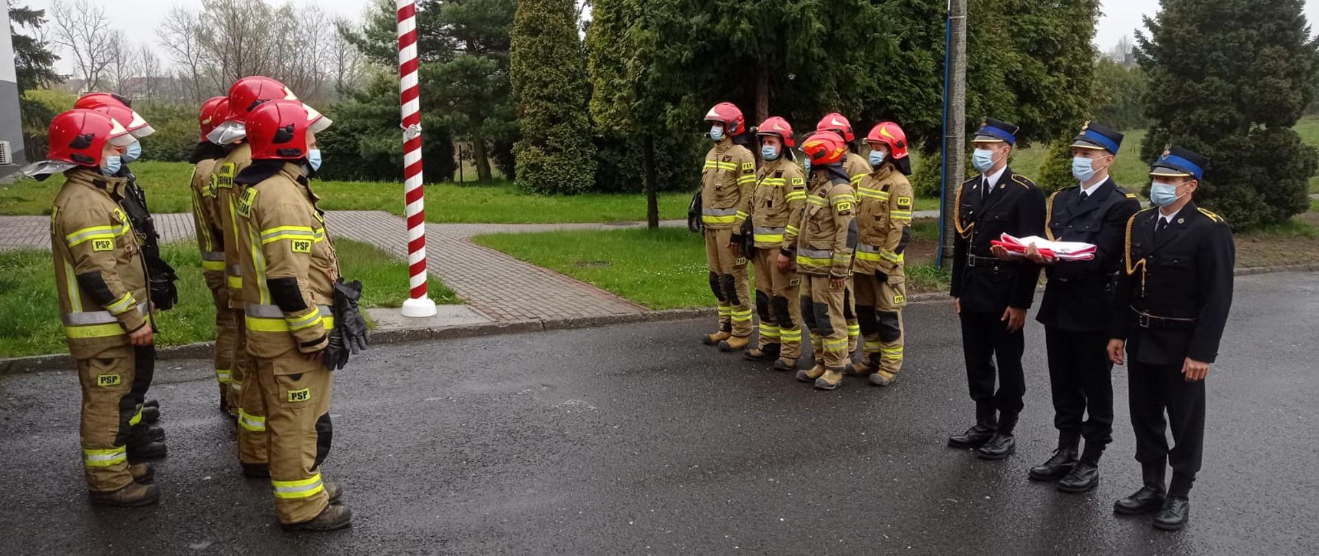 Strażacy stojący na przeciw siebie w mundurach bojowych i hełmach, po prawej stronie trzech strażaków w mundurach wyjściowych jeden trzyma złożoną flagę państwową, w tle maszt flagowy i drzewa. 