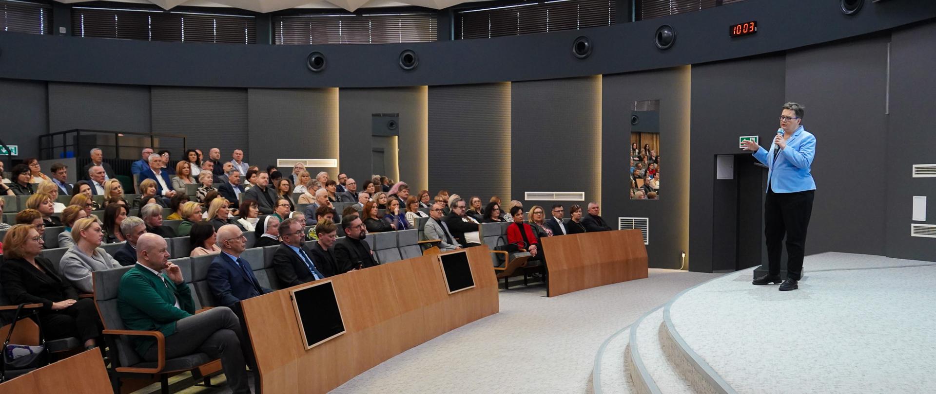 Widok z boku na dużą salę, na ustawionych w rzędy krzesłach siedzą ludzie. Po prawej stronie na podeście stoi wiceminister Lubnauer i mówi do mikrofonu.