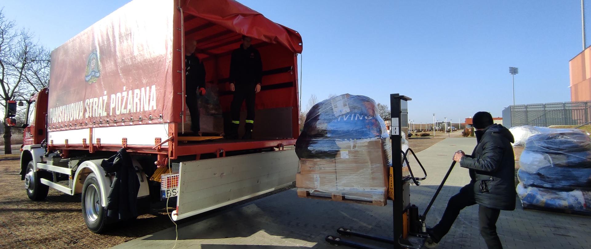 Na zdjęciu widać samochód straży pożarnej na który ładowane są palety z paczkami 