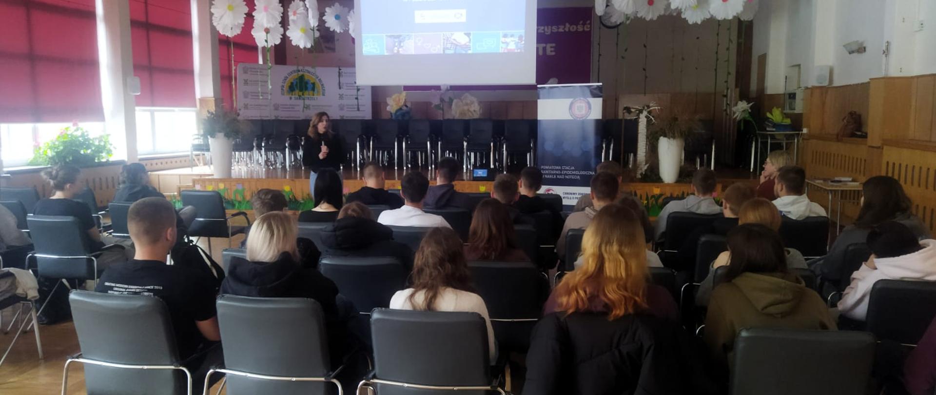 Sala pełna uczniów siedzących na krzesłach, słuchających prelekcji prelegentki na scenie. Na ekranie projekcyjnym widać slajd z logo programu "Młodzi Świadomi".