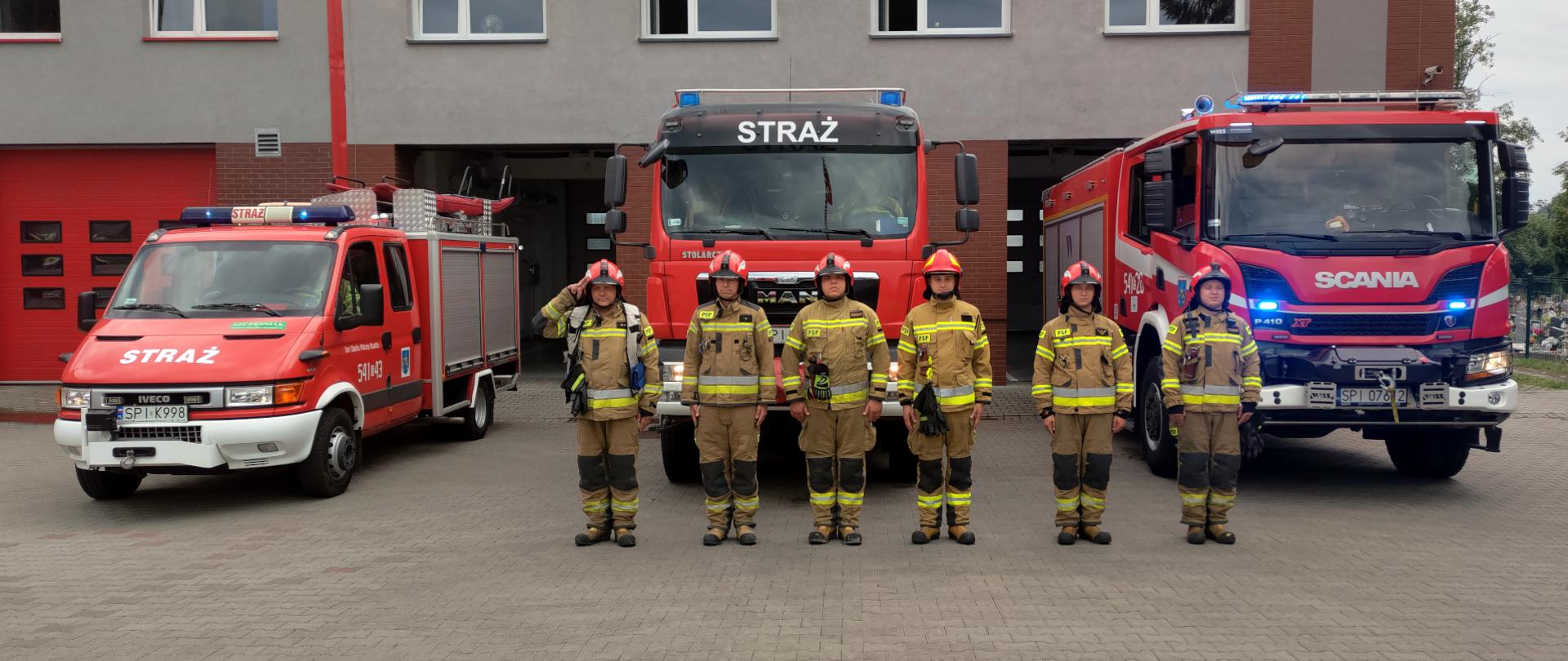 zdjęcie przedstawia trzy samochody ratowniczo gaśnicze z włączonymi sygnałami świetlnymi. Przed samochodami stoi pododdział strażaków w ubraniach specjalnych. Pierwszy z lewej salutuje. 