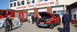 Strażacy i przedstawiciele służb mundurowych oglądają samochody strażackie na placu przed komendą
