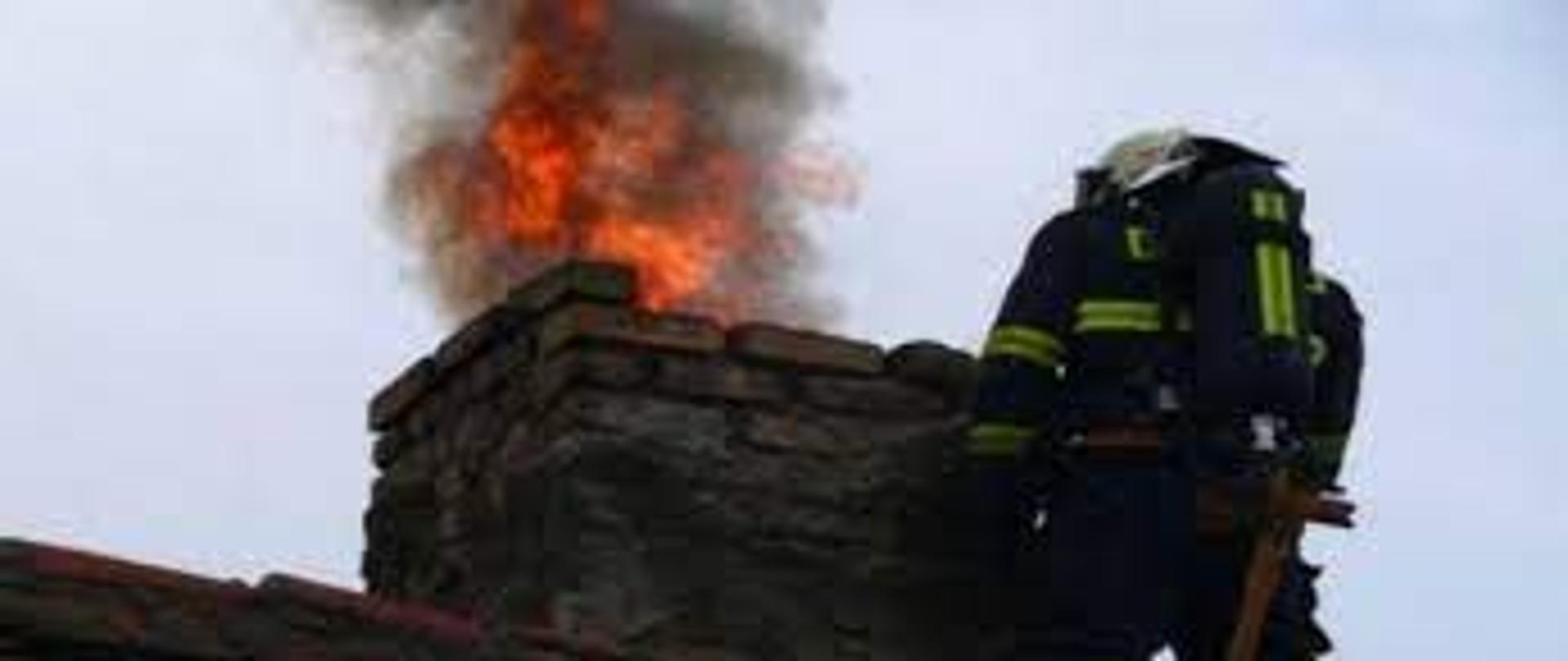 Zdjęcie przedstawia strażaka na dachu budynku podczas pożaru sadzy w kominie. Ze starego uszkodzonego komina wydobywa się ogień i dym.