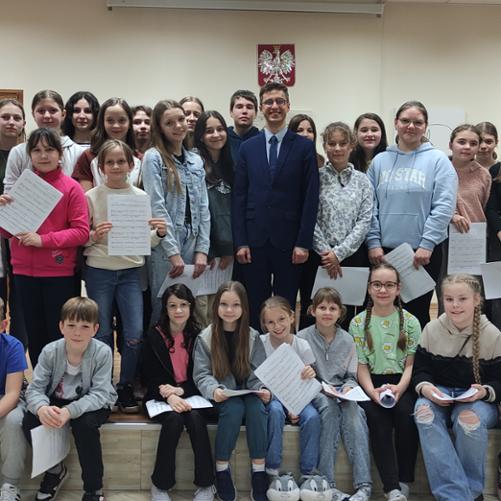 Grupa około 30 osób stoi na scenie. Część z nich trzyma w dłoniach kartki z nutami. W tle na ścianie godło. 