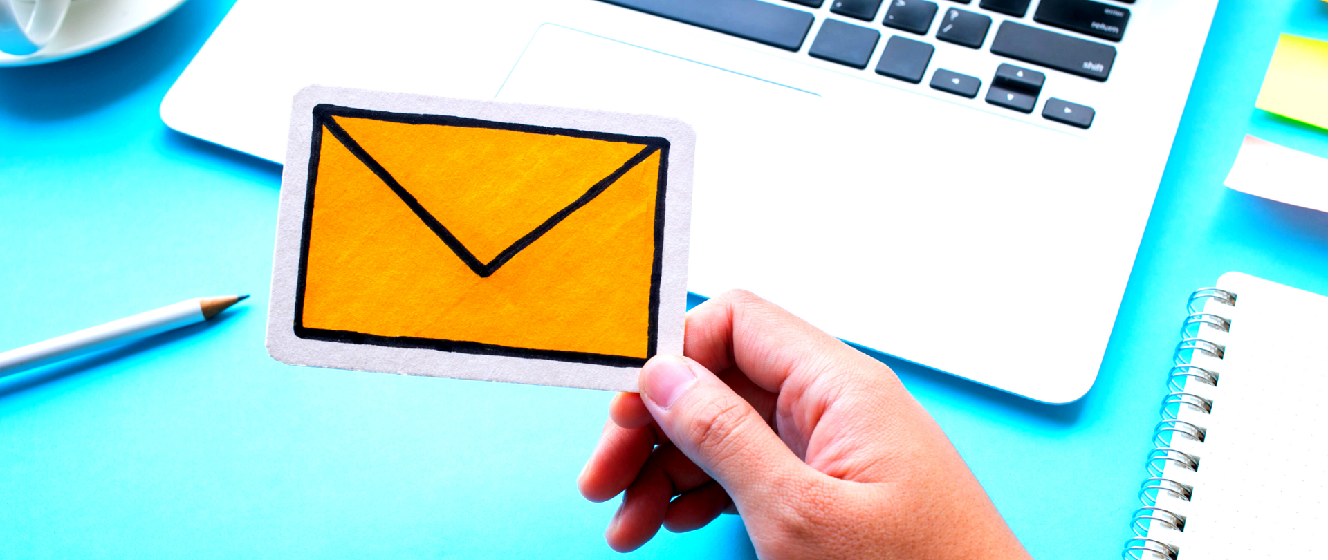 Dłoń trzymająca prostokątny kartonik, na którym narysowana jest żółta koperta - symbolizująca wiadomość elektroniczną. W tle (błękitnym) biały laptop, ołówek i skoroszyt.