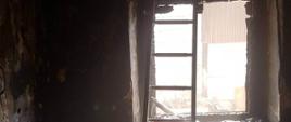 Pożar budynku mieszkalnego. Wnętrze jednego z pomieszczeń, które było objęte ogniem. Widoczne są ślady zniszczeń. W głębi drabina stojąca przy oknie.