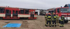 na zdjęciu widać po lewej stronie tramwaje udostępnione do ćwiczeń. Po prawej stronie stoi 7 strażaków PSP ubranych w ubrania specjalne. Kierownik ćwiczeń przekazuje strażakom informacje o sposobie wykonania ćwiczenia.