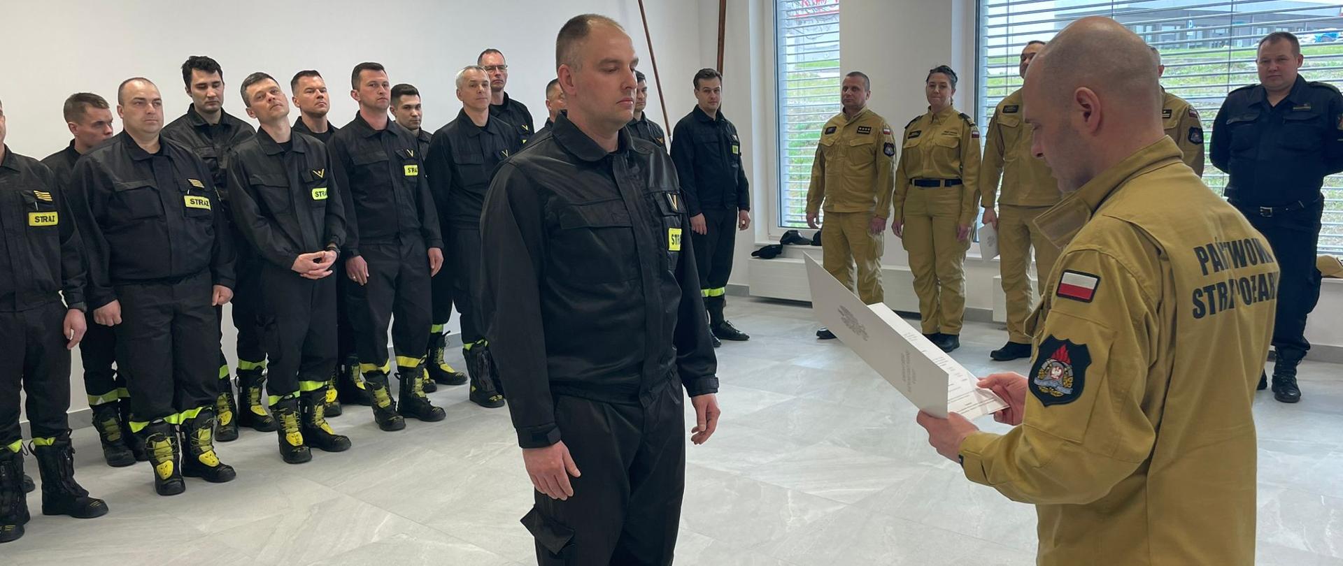 Na pierwszym planie Komendant Miejski wręcza akt mianowania na wyższe stanowisko służbowe podległemu funkcjonariuszowi. W tle stoją w szeregu pozostali funkcjonariusze. 