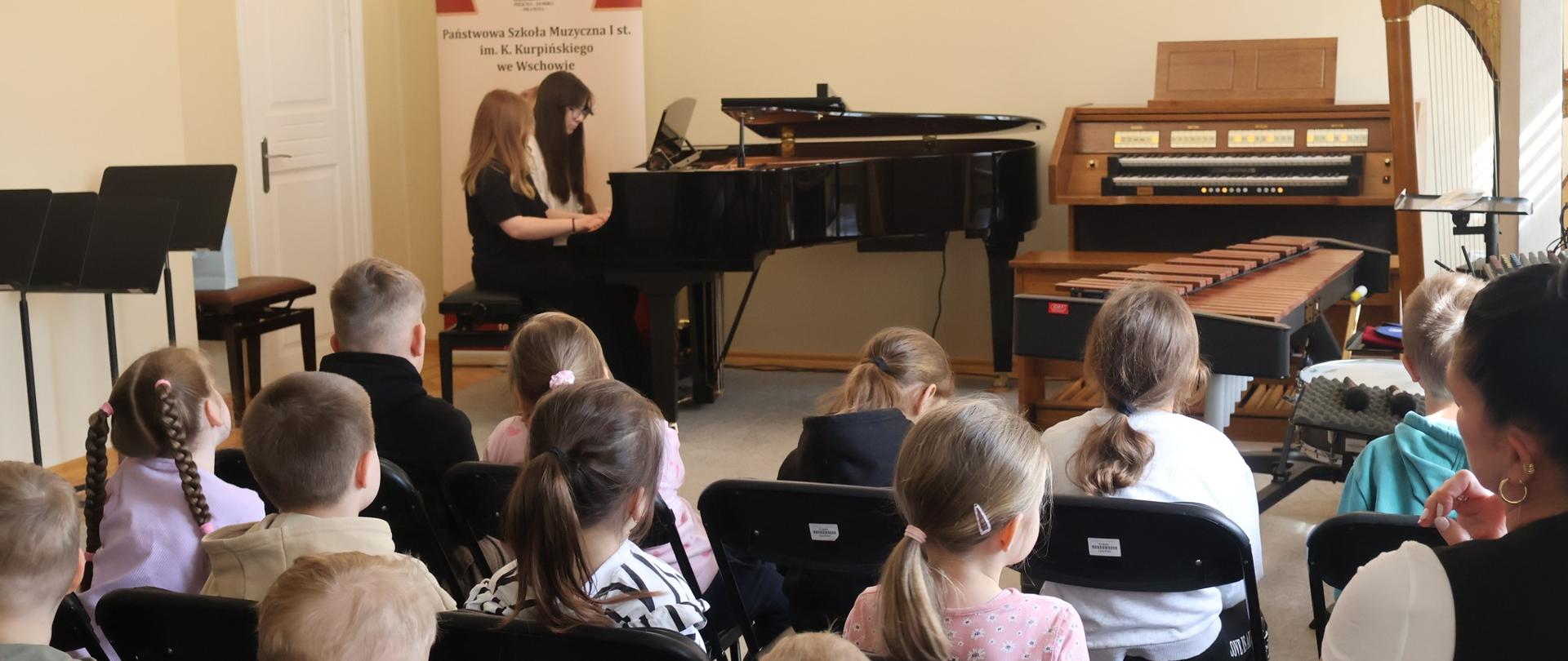 Duet pianistyczny występujący w auli szkoły. Tyłem siedzące dzieci, słuchające występu.