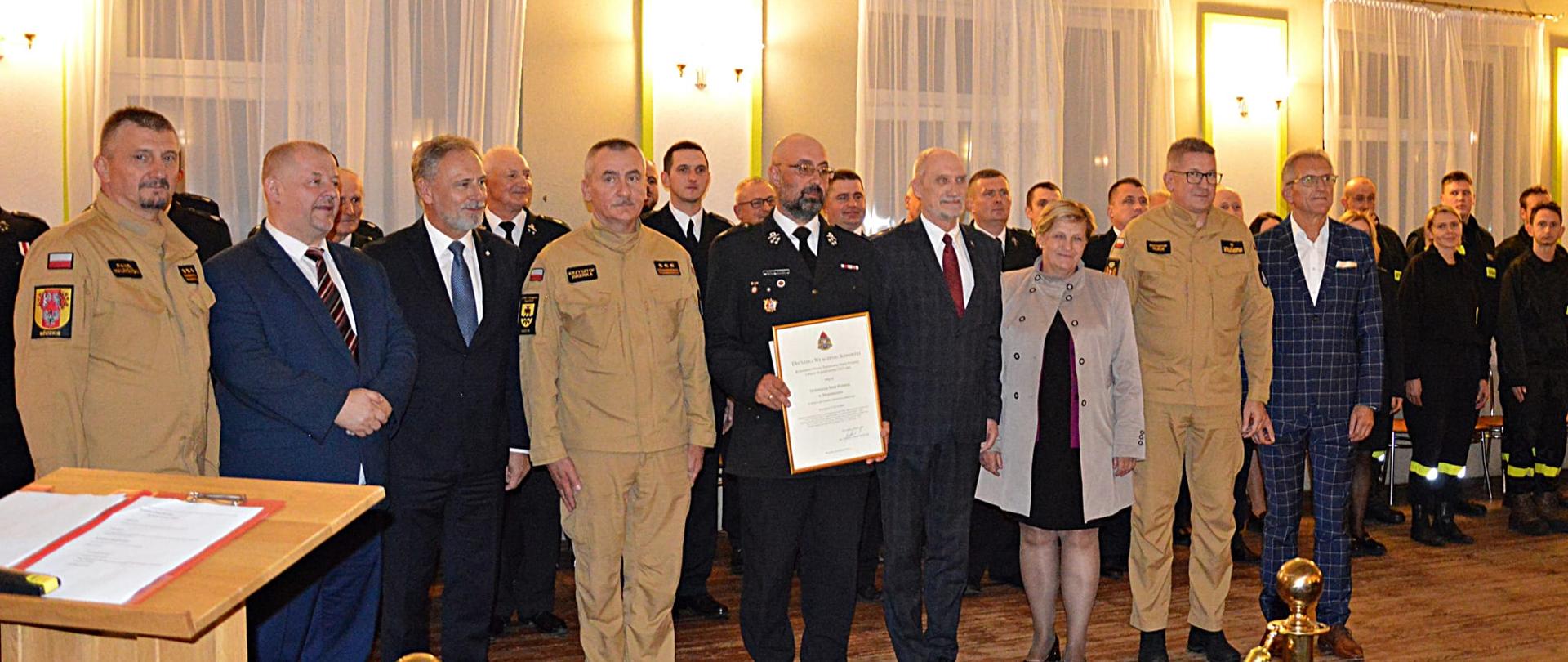 Na zdjęciu widać strażaków, parlamentarzystów i przedstawicieli samorządów stojących w rzędzie. Jeden ze strażaków trzyma w ręku decyzję o włączeniu jednostki do krajowego systemu-ratowniczo gaśniczego. Za nimi stoją druhowie z OSP Strzemeszna.