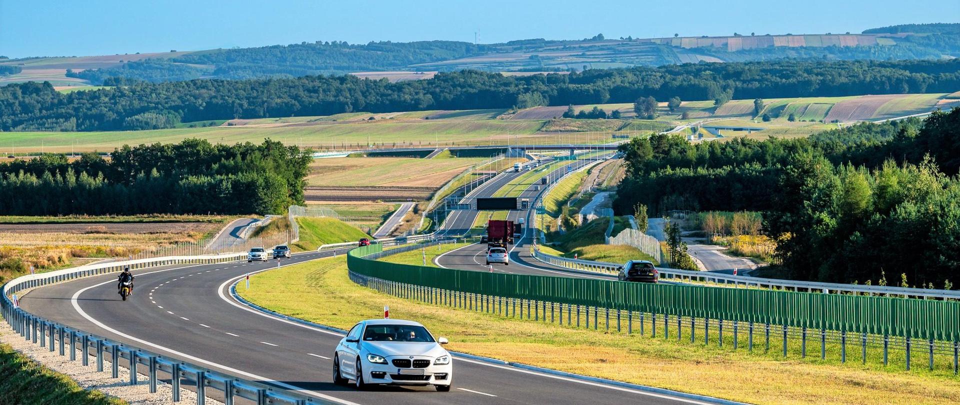 Droga ekspresowa S7 na odcinku Moczydło - Miechów. Dwujezdniowa droga ekspresowa wijąca się między polami i lasami po niewielkich pagórkach. Widoczne pojazdy poruszające się w obu kierunkach.