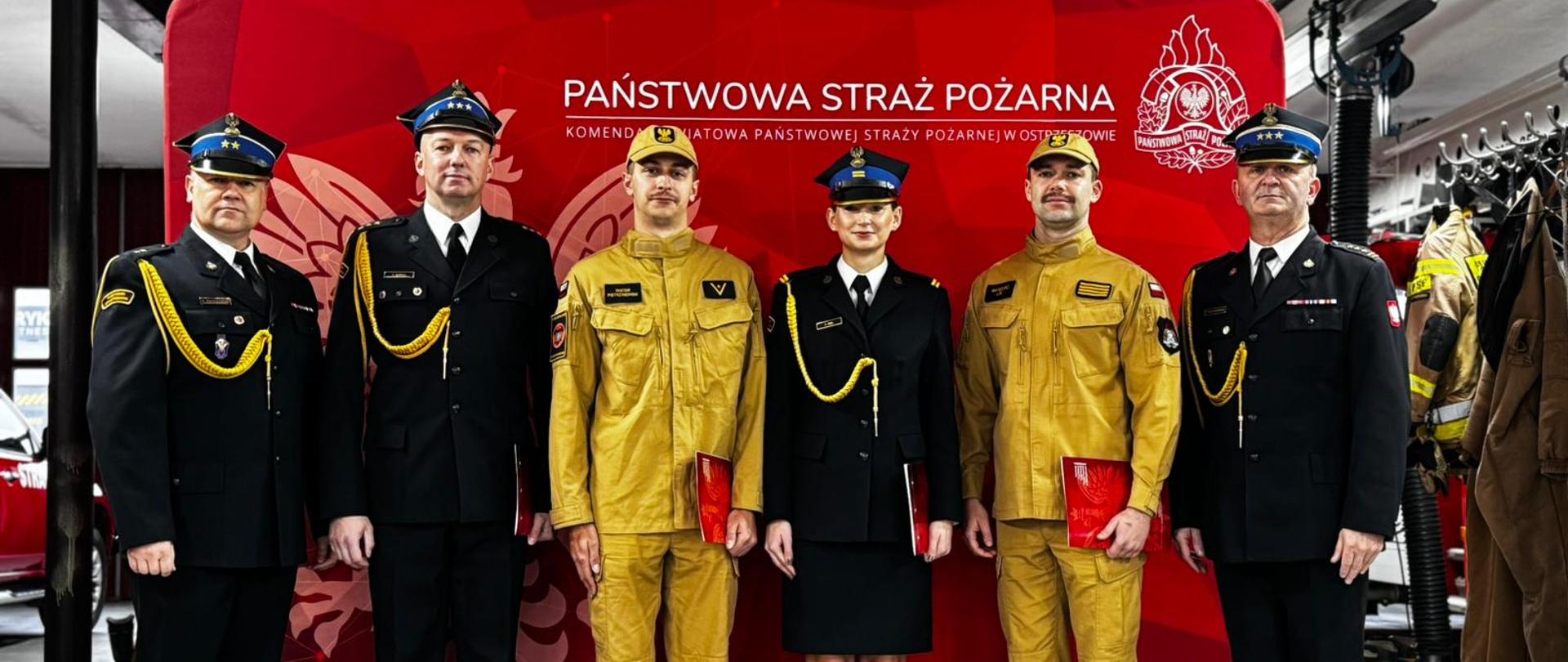 Komendanci oraz wyróżnieni funkcjonariusze na tle czerwonego bannera z logiem Państwowej Straży Pożarnej. W tle samochód strażacki. Zdjęcie zrobione w garażu strażnicy.