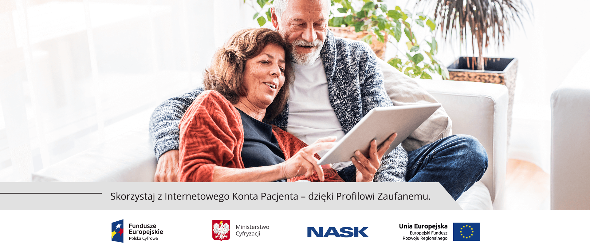 Kobieta i mężczyzna, oboje w średnim wieku. Siedzą na kanapie. Kobieta trzyma tablet. Obok kanapy stoją rośliny w doniczkach