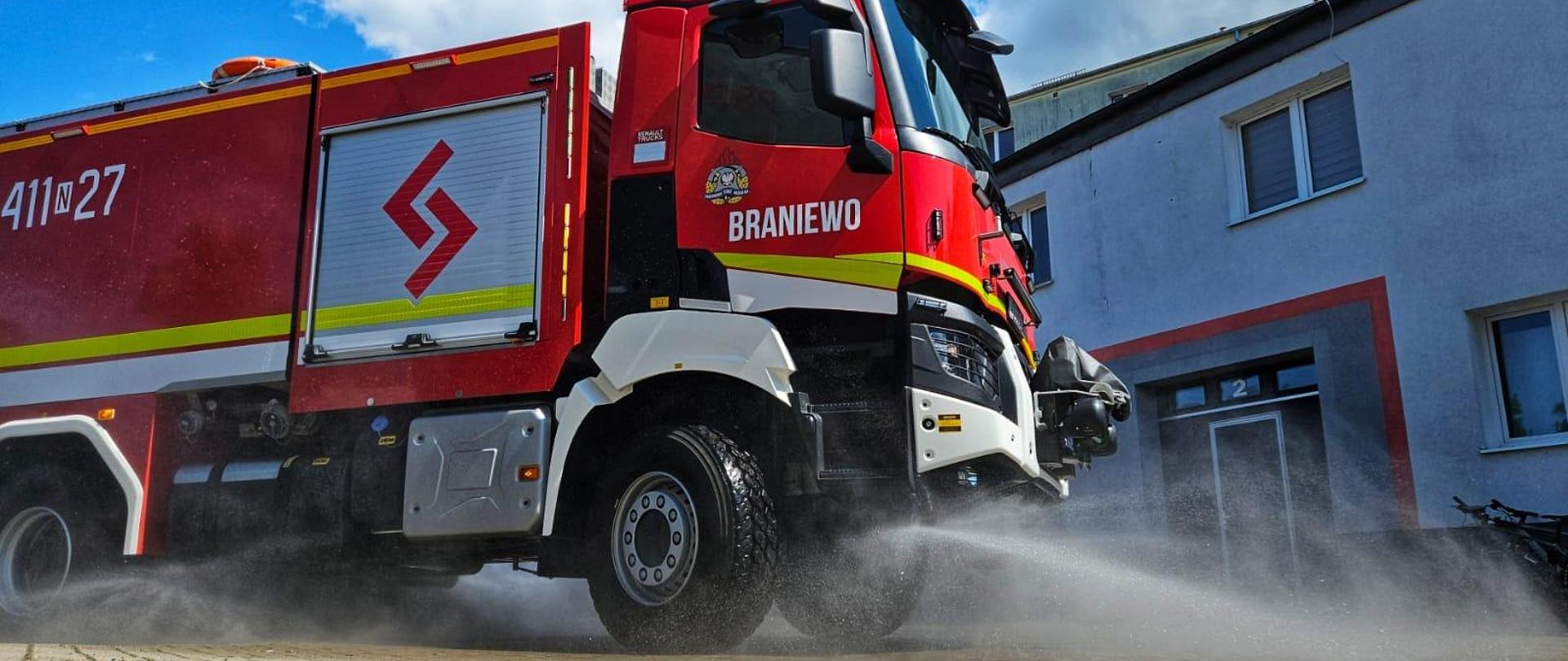 Na zdjęci duży czerwony samochód strażacki. W tle budynek. Nad nim niebieskie niebo.