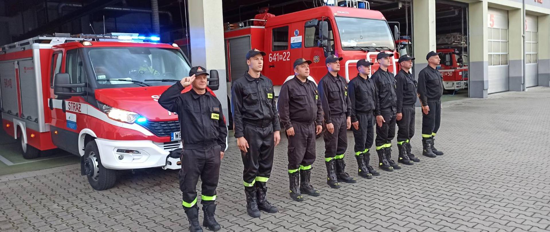 Funkcjonariusze KP PSP w Sokołowie Podlaskim oddali hołd strażakowi, który zmarł na służbie - strażacy stoją na zbiórce przed garażem JRG i samochodami i oddają honory zmarłemu strażakowi.