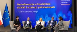 Kilka osób siedzi na fotelach. W tle, na niebieska ściana z napisem: Dezinformacja w kontekście działań instytucji państwowych. KSeF w centrum uwagi.