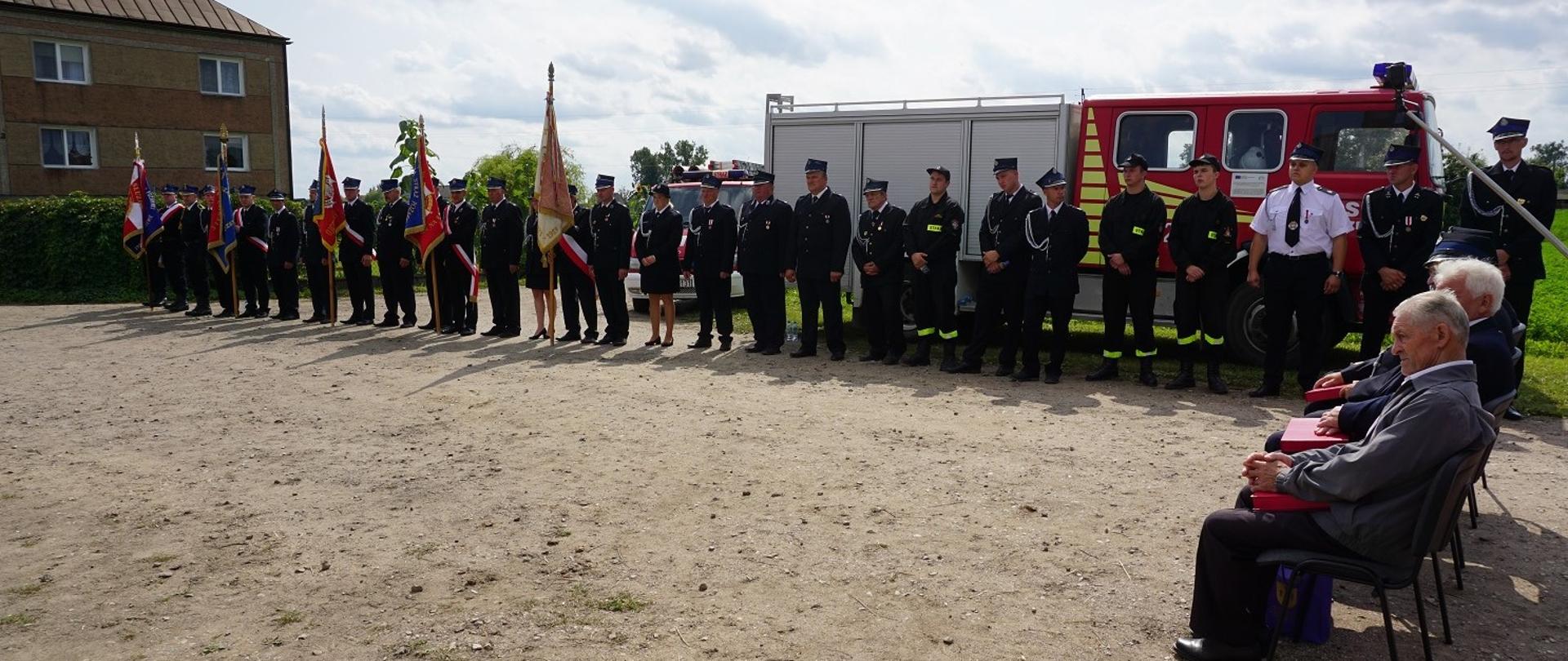 Poczty sztandarowe oraz druhowie OSP stoją w rzędzie. Z prawej na krzesłach siedzą najstarsi druhowie OSP.