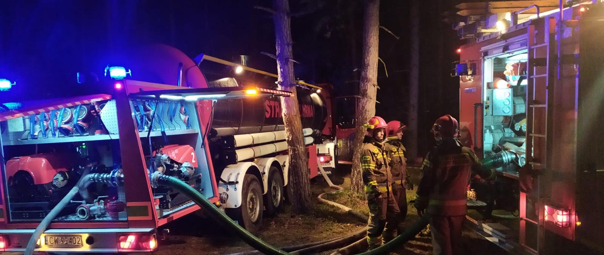 Zdjęcie przedstawia srebrną cysternę strażacką i drugi pojazd gaśniczy stojące w nocy w lesie i połączone liniami wężowymi. Przy sztucznym oświetleniu pracują strażacy.