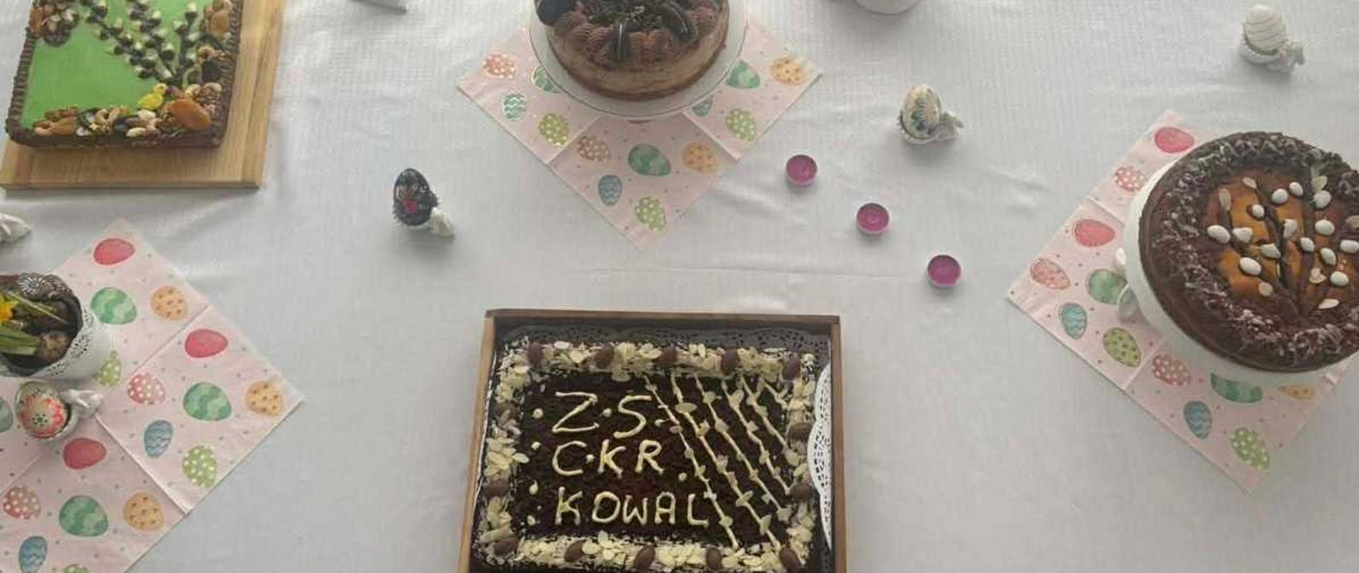 Zdjęcie przedstawia widok z góry na stół przykryty białym obrusem, na którym zaaranżowano wielkanocny poczęstunek. Na stole znajdują się cztery różne ciasta oraz liczne świąteczne dekoracje. Panuje jasna, radosna i odświętna atmosfera.
Wypieki (rozmieszczone na planie koła)
Ciasto prostokątne (na dole): Ciemne ciasto czekoladowe w drewnianej ramce, ozdobione białym lukrem tworzącym wzór kratki. Na środku widnieje staranny napis wykonany jasną masą: „ZS CKR KOWAL”. Boki ciasta udekorowane są małymi, czekoladowymi jajeczkami.
Mazurek (po lewej): Prostokątne ciasto na drewnianej desce. Wierzch pokryty jest jasnozielonym lukrem, na którym ułożono dekorację przypominającą bazia (kotki) z bakalii i orzechów.
Tort (na górze, w centrum): Okrągłe, wysokie ciasto czekoladowe, bogato zdobione na wierzchu ciasteczkami typu Oreo oraz kremowymi rozetkami. Stoi na kolorowej serwetce w pisanki.
Babka lub mazurek okrągły (po prawej): Ciemne, okrągłe ciasto posypane bakaliami i płatkami migdałów, które tworzą wzór kwiatowy lub gałązki.
Dekoracje i detale
Motywy wielkanocne: Na stole rozstawionych jest kilka białych, ceramicznych figurek przedstawiających zające w różnych pozach oraz jajka.
Roślinność: W małej białej doniczce rosną żywe, żółte narcyzy (żonkile).
Akcenty kolorowe: Pod ciastami leżą papierowe serwetki z nadrukiem kolorowych pisanek. Na obrusie rozrzucone są małe, różowe świeczki typu tealight oraz kilka mniejszych, ozdobnych jajeczek.
Tło: Biały obrus ma delikatną strukturę, a cała kompozycja jest bardzo czysta i symetryczna.
