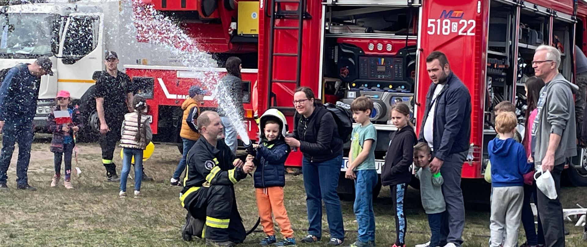 Sopockie Błonia na których odbywa się piknik rodzinny. W tyle widoczny samochód ratowniczo gaśniczy stojący tyłem z otwartymi skrytkami, po jego lewej stronie drabina mechaniczna stójąca tyłem i dalej samochód śmieciarka. Na przodzie widać kolejkę dzieci i doisrosłych ustawioną do strażaka który podaje każdemu prądownicę podpięta do węża i leją wodę