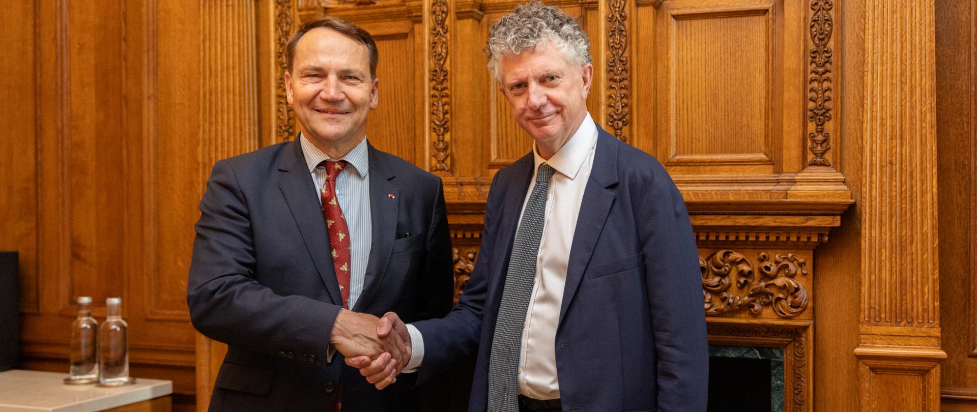 Wicepremier, Minister Spraw Zagranicznych Radosław Sikorski i brytyjski Doradca Narodowy ds. Bezpieczeństwa Jonathan Powell w formalnych garniturach ściska sobie dłonie podczas oficjalnego spotkania. Spotkanie odbywa się w eleganckim wnętrzu z bogato zdobioną drewnianą boazerią i kominkiem w tle. Po lewej stronie widoczny jest stolik z dwiema butelkami wody.