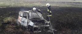 Na zdjęciu rozwinięty wąż gaśniczy, na środku doszczętnie spalony samochód osobowy, przy nim dwóch strażaków.