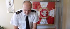 Na zdjęciu widzimy Komendanta Powiatowego Państwowej Straży Pożarnej w Rypinie starszego brygadiera Grzegorza Łydkowskiego. Znajduje się On w budynku Komendy Powiatowej, w gabinecie Komendanta. Pan starszy brygadier Grzegorz Łydkowski składa podpis na akcie ślubowania, które złożyła Pani strażak Justyna Pilśniak. W tle, w gablocie znajduje się sztandar Komendy Powiatowej Państwowej Straży Pożarnej w Rypinie.