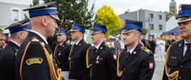 Oficer Państwowej Straży Pożarnej wręcza dokumenty gratulacyjne strażakom w mundurach galowych podczas oficjalnej ceremonii na placu.