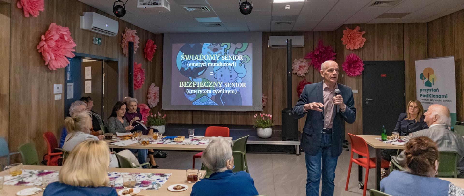 Pan Leszek Szreder przemawia podczas spotkania Świadomy Senior - Bezpieczny Senior w Piasecznie. Uczestnicy spotkania siedzą przy stołach. W tle, na ścianie wyświetlana jest prezentacja do wykładu.