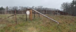 Na zdjęciu widać teren trawiasty, w oddali ogrodzenie z siatki zamontowane na drewnianych kołkach z zamontowana kładką opadającą po obu stronach ogrodzenia. W oddali widać ogrodzenie drewniane.