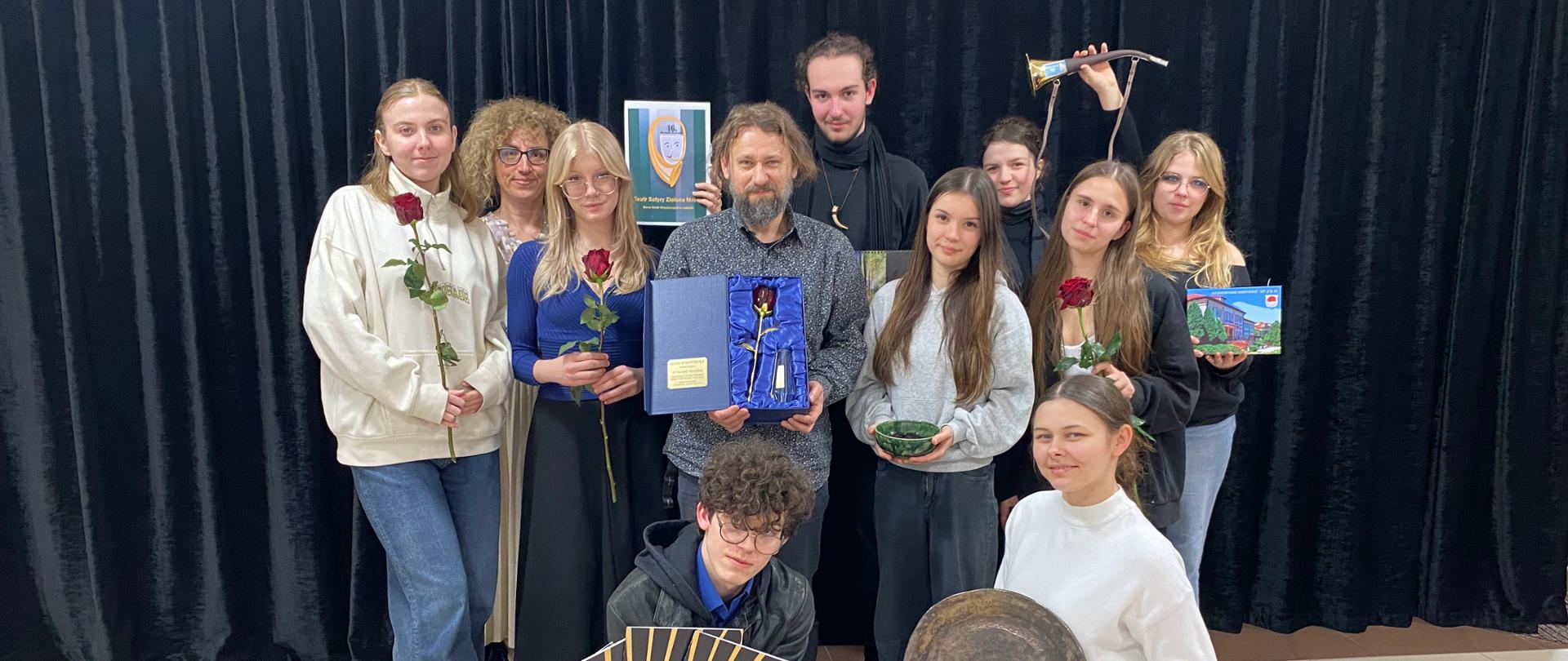 Na tle czarnej kurtyny do zdjęcia pozuje 11 - osobowa grupa. Jeden chłopiec na dole przyklęka i trzyma w dłoniach kilka żółtobrązowych teczek, obok niego dziewczyna trzyma duży szary talerz. W górnym rzędzie pośrodku mężczyzna trzyma metalową różę w niebieskim etui. Po prawej stronie dziewczyna trzyma zieloną miseczkę, kolejna czerwoną różę, kolejna zdjęcie budowli z Horyńca. stojący za nimi dziewczyna i chłopiec trzymają róg. Po lewej stronie stoją dwie dziewczyny z czerwonymi różami, za nim istoi kobieta trzymająca zielono-niebiesko-żółty dyplom z napisem Teatr Satyry Zielona Mrówa