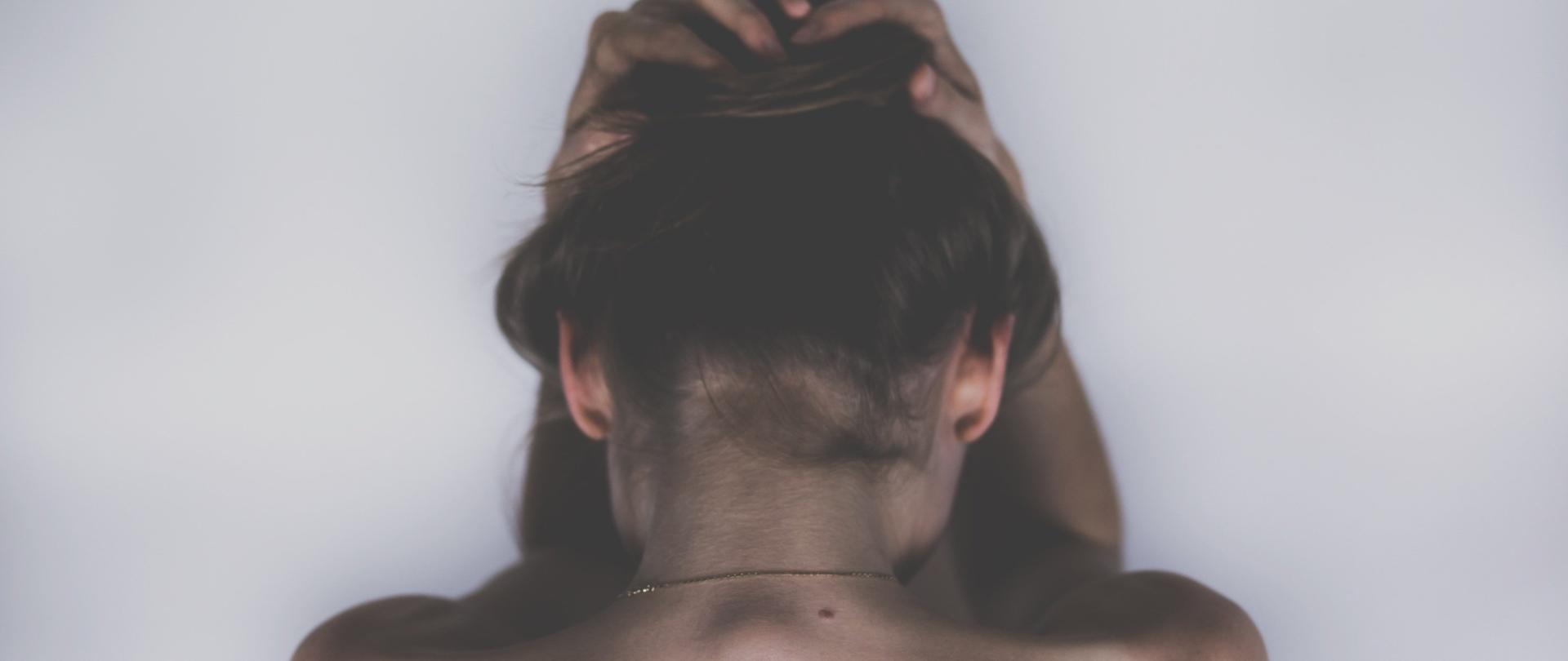 Zdjęcie przedstawia górną część tułowia i głowę kobiety od tyłu. Chwyta się ona w geście bezradności obiema rękoma za głowę. Co symbolizuje niemoc i niepokój wewnętrzny.