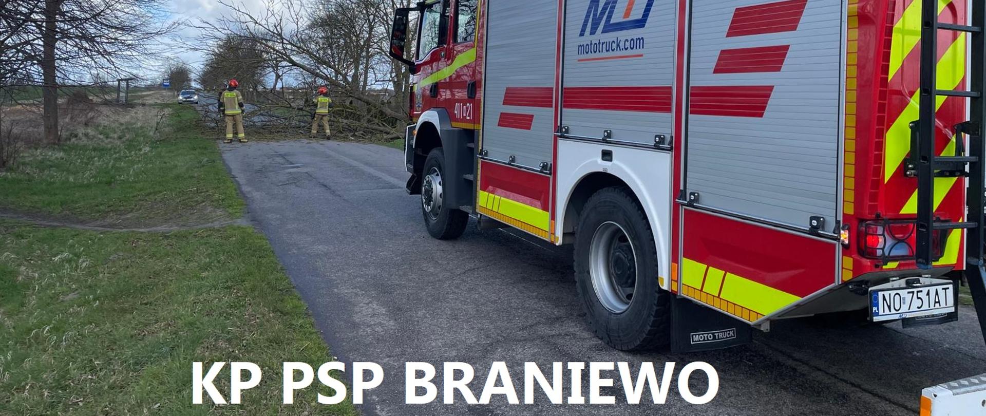 Z lewej strony stoi duży czerwony samochód strażacki. Dalej w oddali drzewo w poprzek drogi przy nim strażacy. Niebieskie niebo.