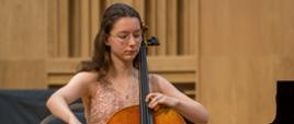 Na scenie, na krześle zasiada młoda dziewczyna. Ma na sobie piękną, wieczorową suknię. Gra na wiolonczeli.