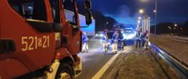 Na pierwszym planie (od lewej): samochód ratowniczo-gaśniczy straży pożarnej. W tle: strażacy dogaszają pożar komory silnika samochodu osobowego. Miejsce zdarzenia zabezpiecza oznakowany furgon śląskiej Inspekcji Transportu Drogowego z włączonymi światłami niebieskimi uprzywilejowania w ruchu drogowym.