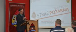Narada roczna. Świetlica Komendy Powiatowej PSP w Rawiczu. Przy mównicy, na tle ścianki promocyjnej z logo PSP przemawia prezes ZOP ZOSP RP w Rawiczu. Mężczyzna ubrany jest w garnitur kratę. Z prawej strony znajduje się ekran, na którym wyświetlana jest prezentacja multimedialna. 