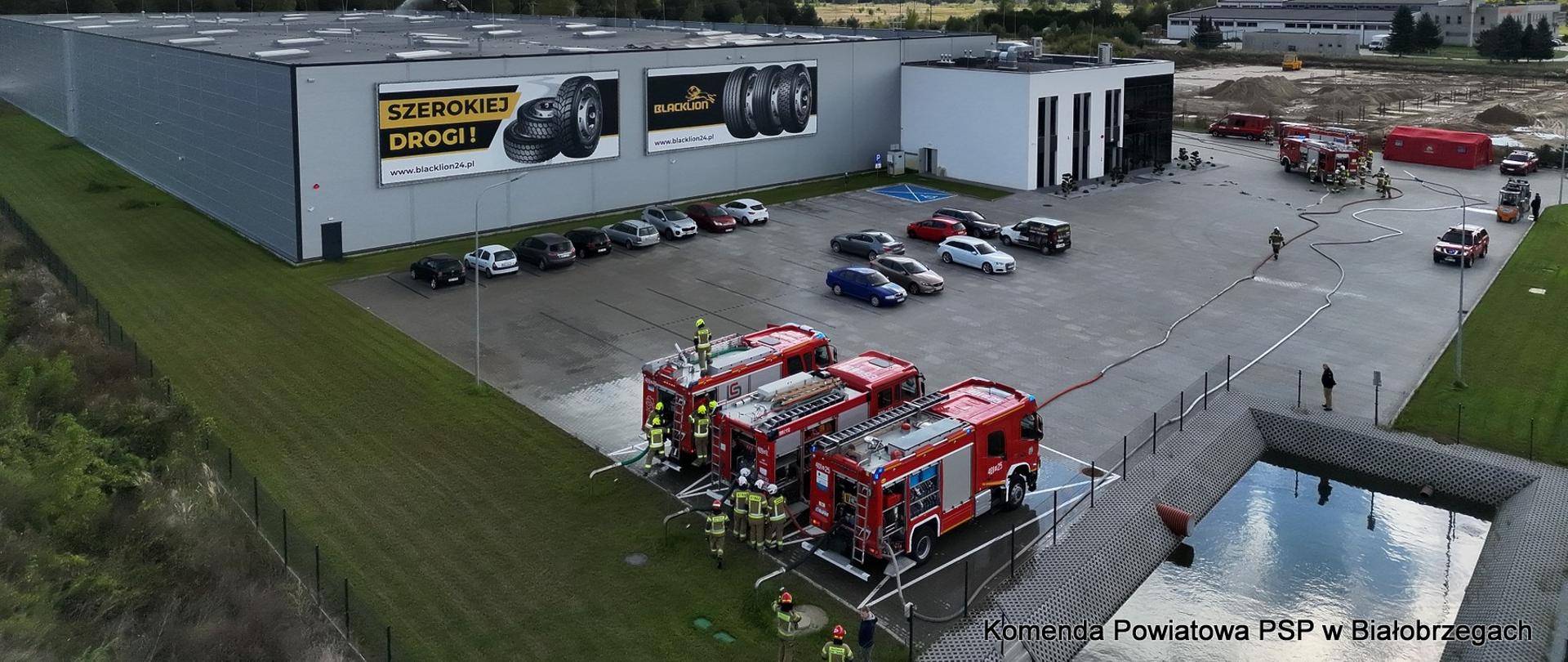 Widok z góry na dużą halę magazynową z napisem "SZEROKIEJ DROGI!". Na parkingu przed halą stoją trzy samochody strażackie z rozłożonymi wężami oraz zaparkowane auta osobowe. Po prawej stronie widać zbiornik wodny. Strażacy prowadzą działania na placu.