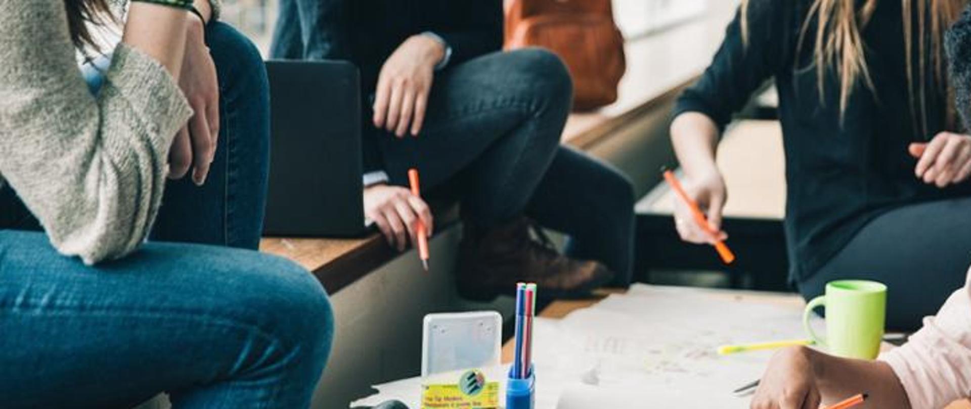 Na zdjęciu są widoczne cztery siedzące osoby, które mają przed sobą kartki papieru a w dłoniach trzymają długopisy. Na biurku oprócz notatek znajdują się również kolorowe długopisy oraz kubek.