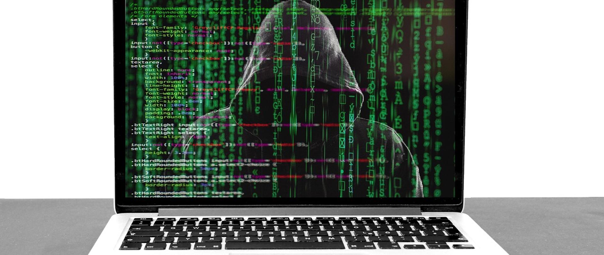 Zdjęcie laptopa. Na ekranie laptopa zamaskowana osoba. W tle kody systemowe.