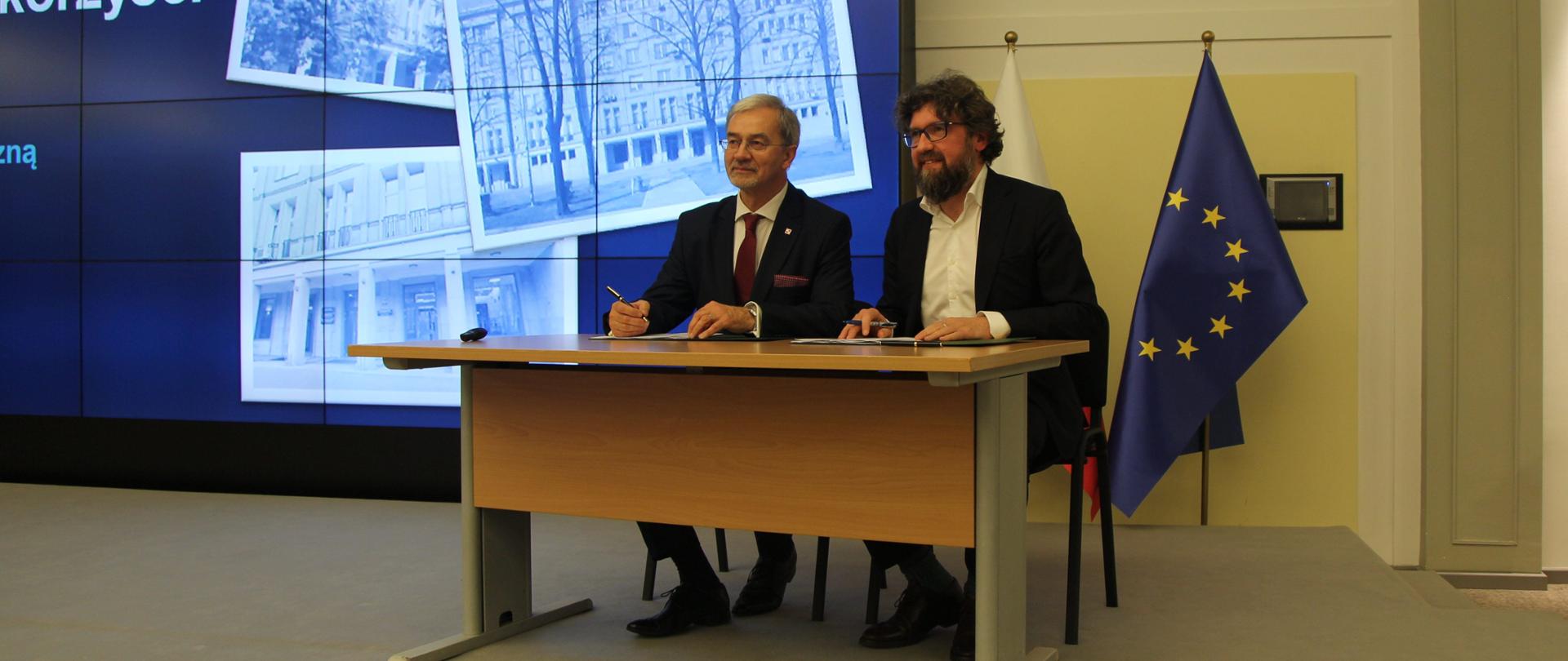 Na sali konferencyjnej na scenie przy stole siedzą od lewej Jerzy Kwieciński oraz Piotr Woźny. Przed nimi leżą dokumenty, które podpisują.