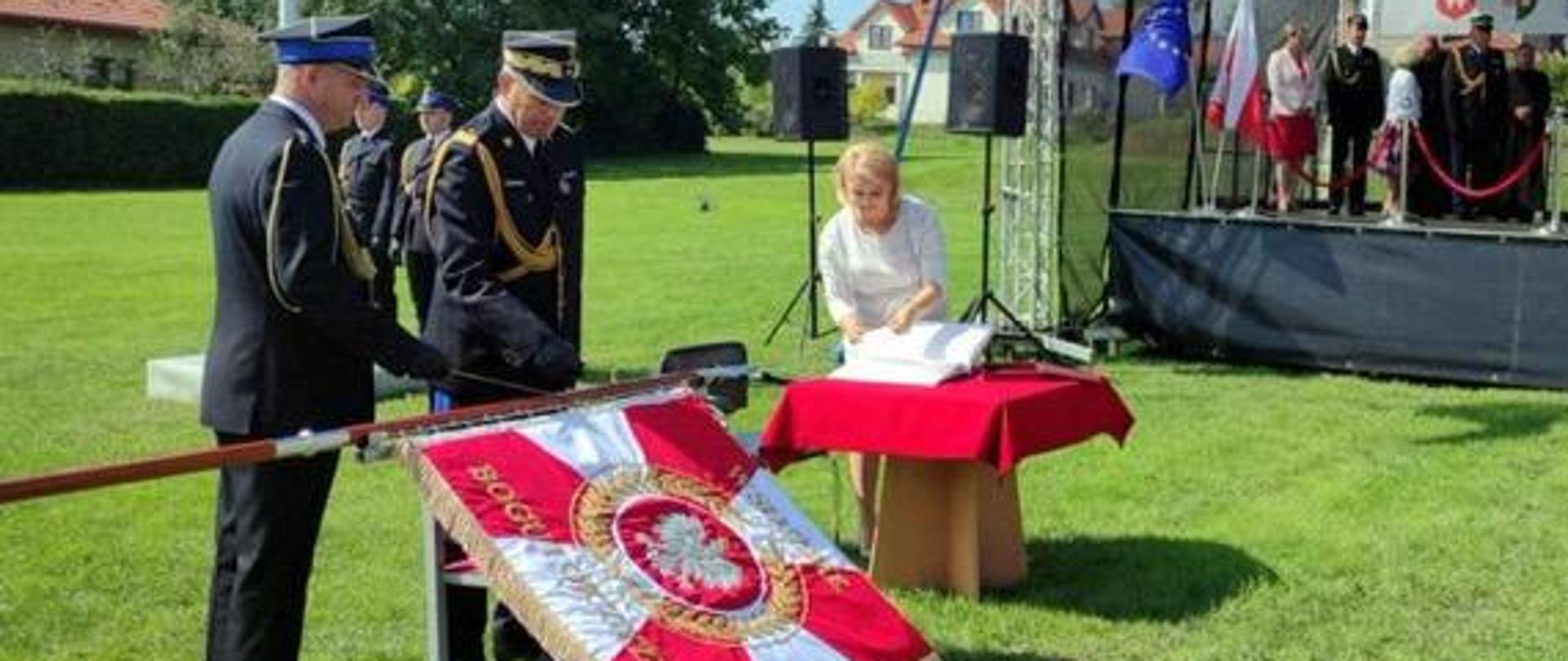 Zastępca Komendanta Głównego PSP i jeszcze jeden funkcjonariusz stoją przy sztandarze, obok kobieta przy stoliku nakrytym czerwonym obrusem otwiera księgę pamiątkową, w tle scena i inni uczestnicy obchodów