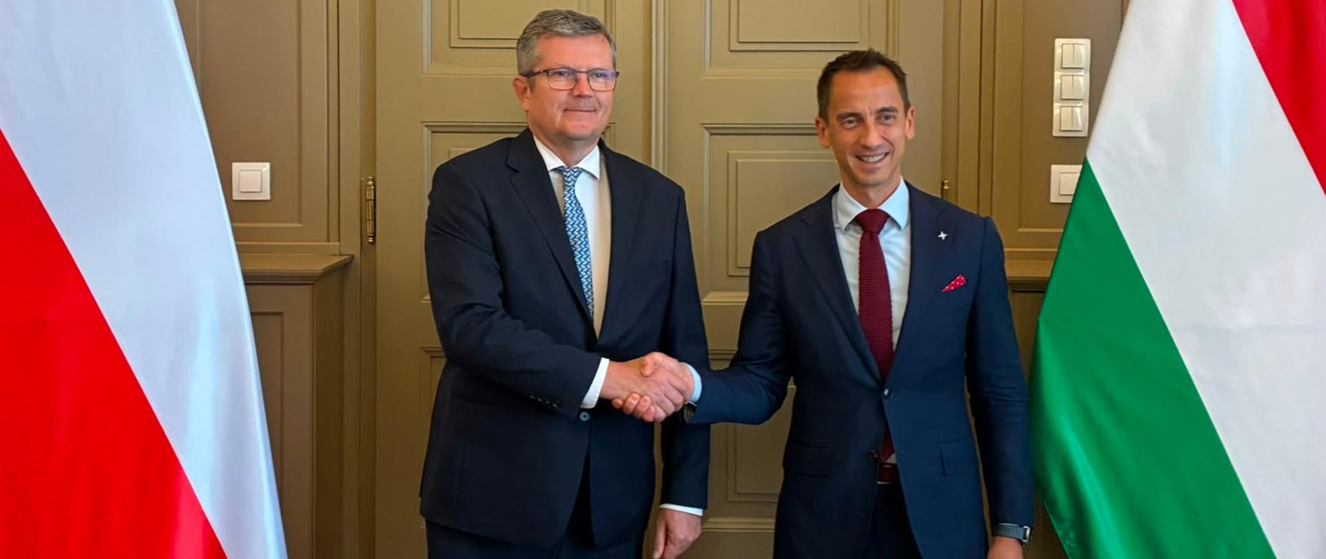 Deputy Ministers of Foreign Affairs of Poland and Hungary shake hands for a photo during an official meeting. They stand in front of ornate double doors between the flags of Poland and Hungary. Both are facing the camera and smiling.