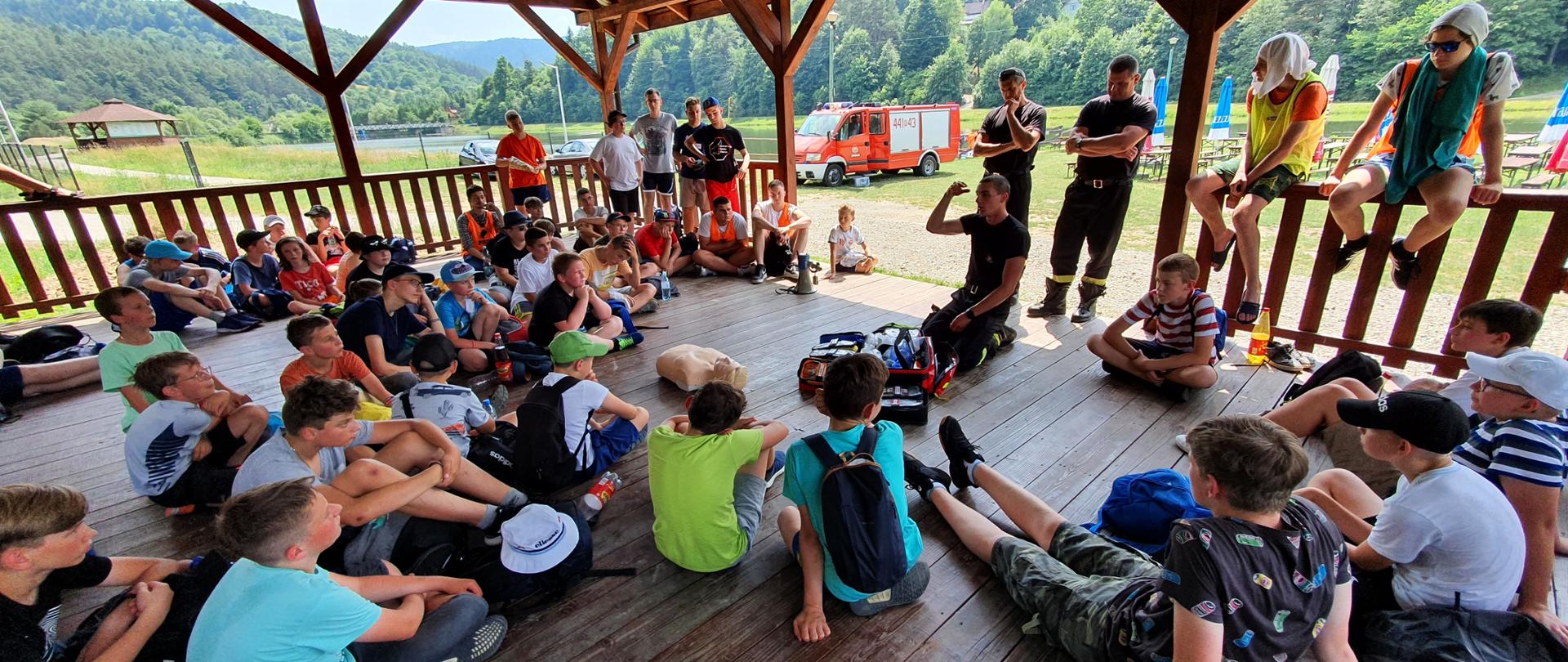 Dzieci i młodzież siedzą w altanie, strażak tłumaczy im zasady udzielania pierwszej pomocy 
