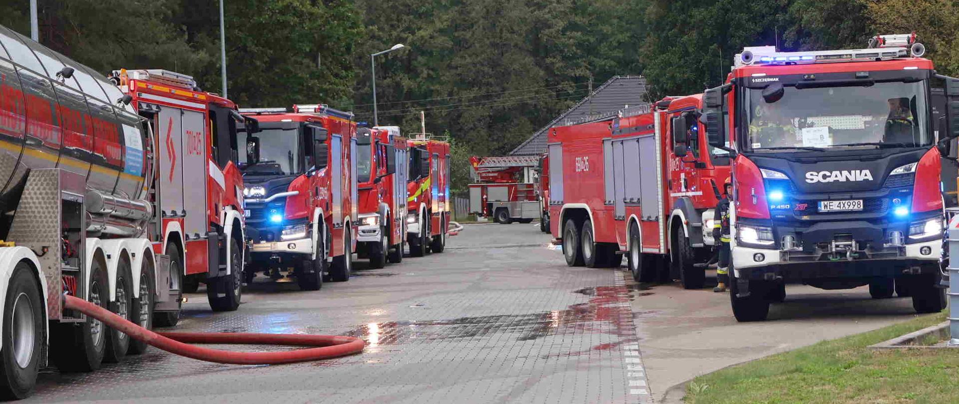 Zdjęcie wykonane podczas ćwiczeń na zakładzie w pionkach. Na zdjęciu widać rząd pojazdów pożarniczych po dwóch stronach drogi.