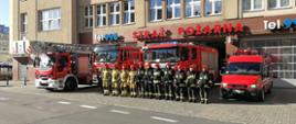 Na zdjęciu można zobaczyć strażaków stojących na baczność przed jednostką ratowniczo-gaśniczą. Za ich plecami stoją równo zaparkowane pojazdy strażackie.