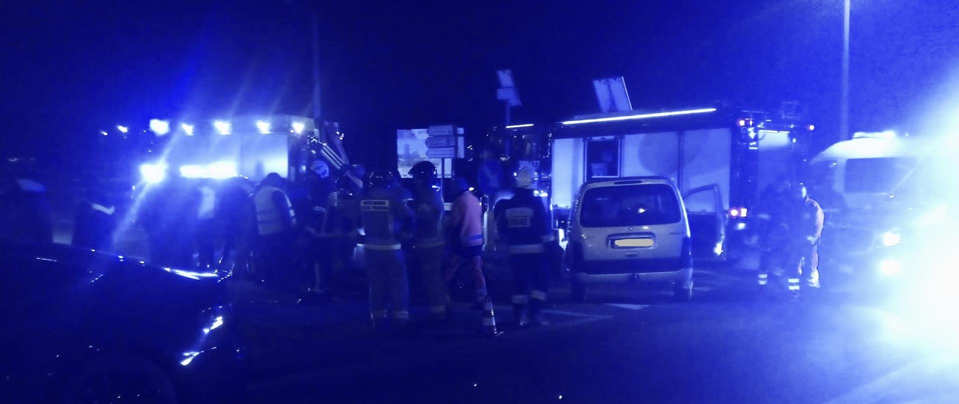 Zdjęcie przedstawia ulicę na której doszło do wypadku komunikacyjnego. W centralnym punkcie grupa ratowników, z prawej i lewej strony dwa z uszkodzonych aut. W tle dwa samochody pożarnicze.